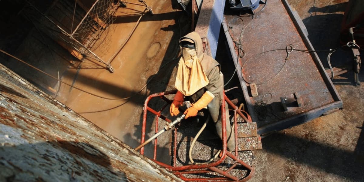Worker performing high-pressure cleaning on a rusted marine vessel surface